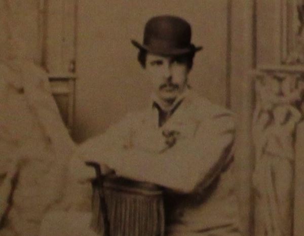 photo of a man posing casually on a chair turned backwards, wearing a bowler hat and white suit